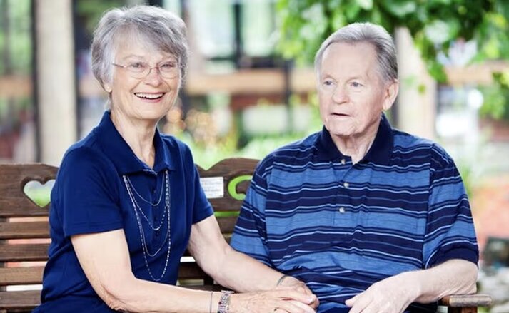 A happy, smiling elderly couple sit on a park bench together at Traditions of Westfield in Westfield, IN